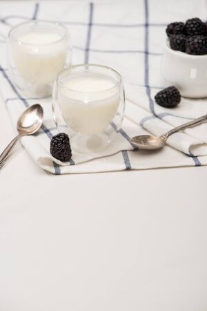 Selective focus of glasses of homemade yogurt, teaspoons and sugar bowl with blackberries on cloth on whiteの写真素材
