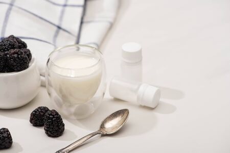 Selective focus of glass of yogurt, teaspoon, containers with starter cultures and sugar bowl with blackberries on whiteの写真素材