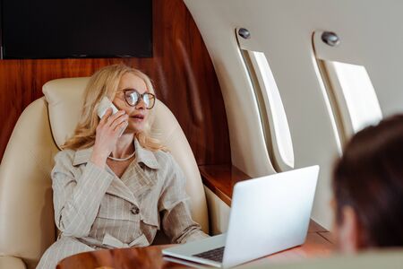 Selective focus of businesswoman talking on smartphone near laptop and businessman in plane