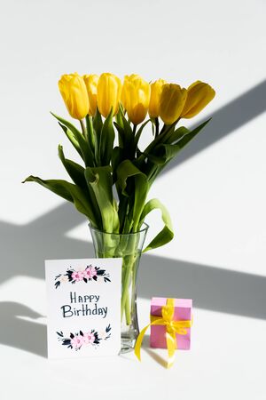 sunlight on yellow tulips in vase near gift box and greeting card with happy birthday lettering on whiteの写真素材