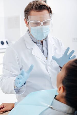 selective focus of dentist in face shield, medical mask and latex gloves gesturing near african american womanの写真素材