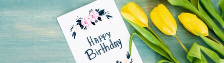 panoramic shot of greeting card with happy birthday lettering near yellow tulips on blue textured...