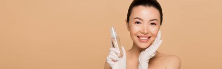 panoramic shot of cheerful nude asian woman in latex gloves holding antiseptic spray isolated on beigeの写真素材