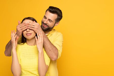 Smiling man covering eyes to smiling girlfriend on yellow backgroundの写真素材