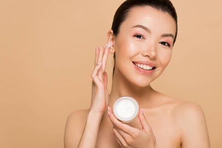beautiful nude asian woman applying cosmetic cream on face isolated on beigeの写真素材
