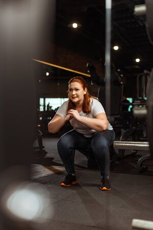 selective focus of overweight girl squatting with clenched hands in gymの写真素材