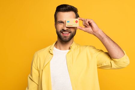 Smiling man covering eye with model of credit card and looking at camera isolated on yellow