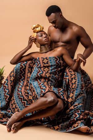 sexy naked tribal afro woman covered in blanket posing near man on beigeの写真素材