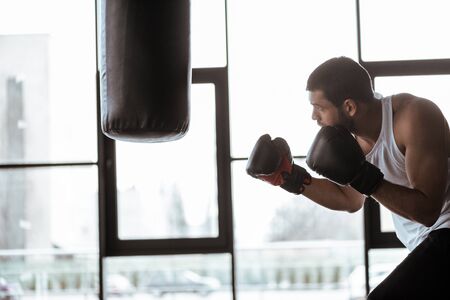 side view of handsome sportsman in boxing gloves training with punching bagの写真素材