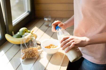 cropped view of woman holding bottle with milk near cornflakes in bowl and fruitsの写真素材