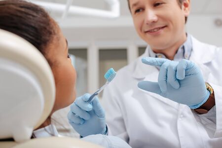 selective focus of happy dentist gesturing near african american woman while holding toothbrushの写真素材