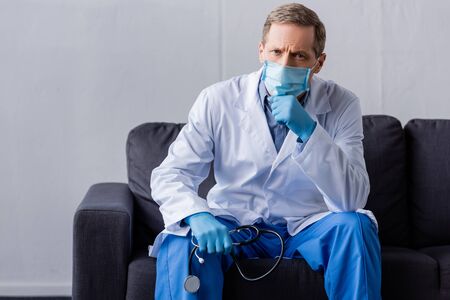 mature doctor in medical mask holding stethoscope while sitting on sofa