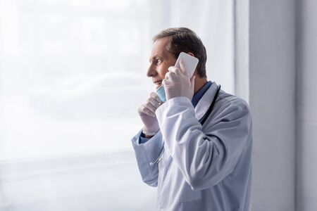 happy and mature doctor in white coat talking on smartphone near window