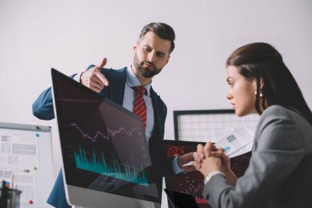 Selective focus of data analyst with papers pointing on graphs on computer monitor to colleague in...