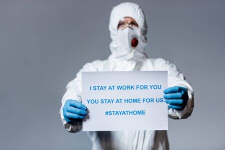 selective focus of mature doctor in hazmat suit holding placard with i stay at work for you, you stay at home for us lettering isolated on greyの写真素材