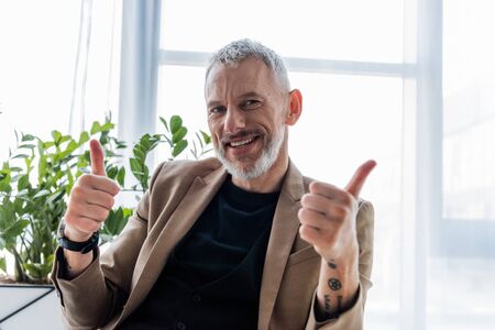 happy and tattooed businessman showing thumbs up while looking at camera