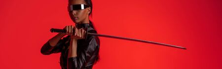 attractive futuristic african american woman in glasses with sword isolated on red, panoramic shotの写真素材