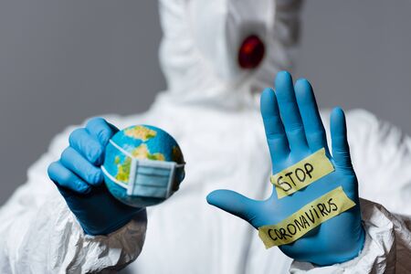selective focus of man in hazmat suit holding globe and showing hand with stop coronavirus lettering isolated on greyの写真素材