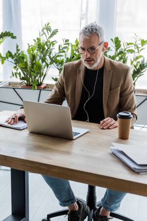 bearded businessman in glasses and earphones looking at laptop near paper cup on table