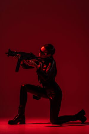 futuristic african american woman in glasses aiming assault rifle on red backgroundの写真素材