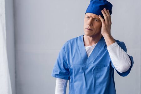 exhausted and middle aged doctor in blue scrub hat touching head on grey