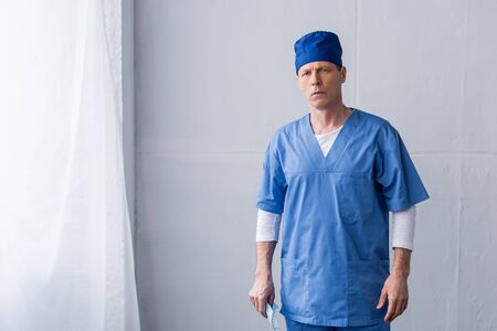 mature doctor in blue scrub hat holding medical mask and standing on grey