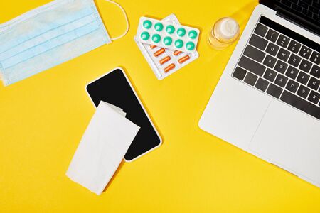 top view of laptop and smartphone with blank screen near napkin, pills, medical mask and bottle with hand sanitizer on yellowの写真素材