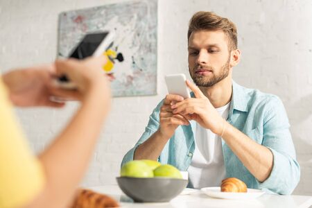 Selective focus of man using smartphone near girlfriend during breakfast in kitchenの写真素材