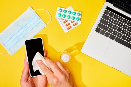 top view of man holding napkin near smartphone with blank screen, laptop, pills, hand sanitizer and medical mask on yellowの写真素材