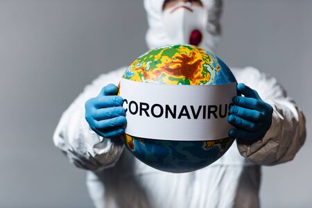 selective focus of man in hazmat suit and medical mask holding globe and paper with coronavirus lettering isolated on greyの写真素材