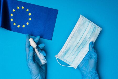 Top view of doctor holding medical mask and bottle of hand sanitizer near flag of european unity on blue backgroundの写真素材
