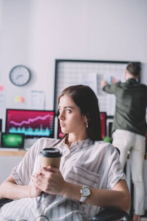 Selective focus of computer systems analyst holding paper cup while colleague in officeの写真素材