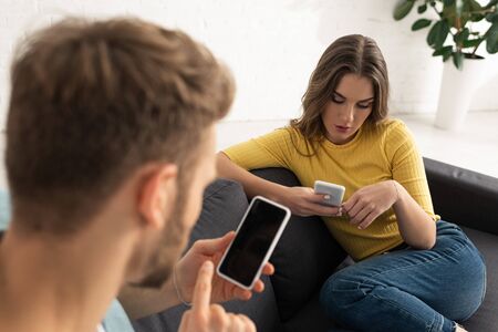 Selective focus of young woman using smartphone near boyfriend on sofa