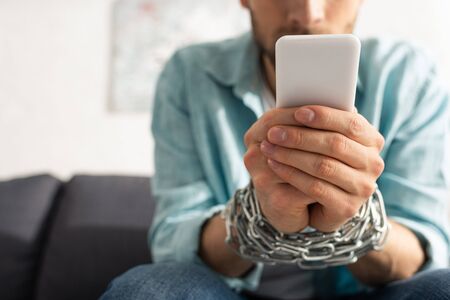 Selective focus of man with tied hands with chain using smartphone on couchの写真素材