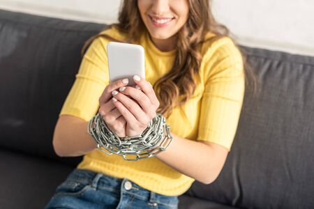 Selective focus of smiling girl with chain around hands holding smartphone on sofaの写真素材