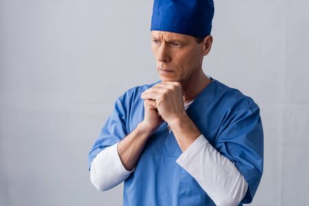 middle aged doctor in scrub hat standing with clenched hands while thinking on greyの写真素材