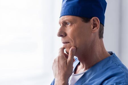 pensive middle aged doctor in scrub hat touching face while thinking on white