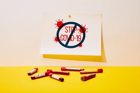 paper with stop covid-19 lettering near test tubes with blood samples on white and yellowの写真素材