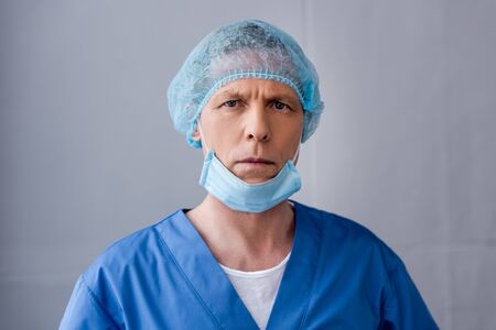 mature doctor in blue medical cap and mask looking at camera on grey