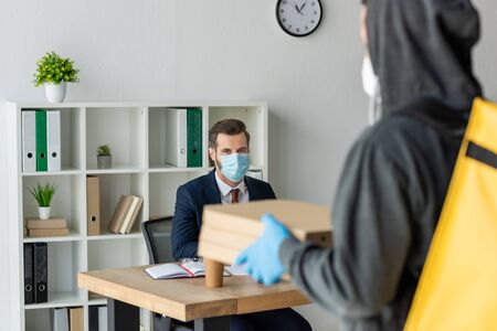 selective focus of food delivery man holding pizza boxes near businessman in medical mask sitting at workplace in officeの写真素材
