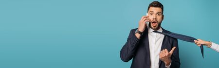 cropped view of woman pulling tie of shocked businessman talking on smartphone and pointing with...