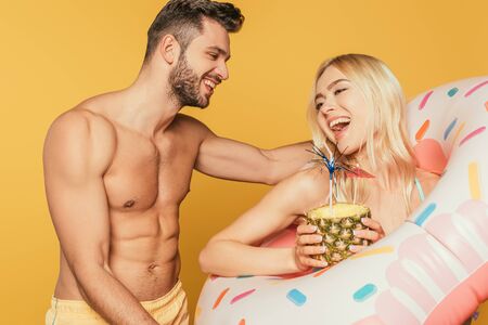 smiling muscular man touching excited girl in swim ring holding pineapple with cocktail isolated on yellowの写真素材