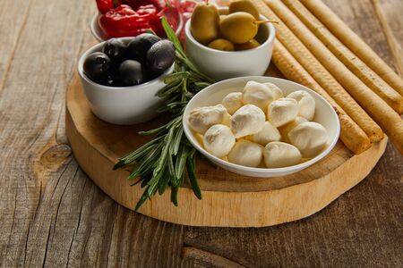 Round board with rosemary, breadsticks and bowls with antipasto ingredients on wooden backgroundの写真素材