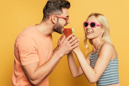 cheerful blonde girl feeding handsome boyfriend with juicy watermelon on yellow backgroundの写真素材