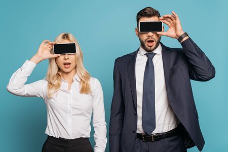 surprised businessman and businesswoman covering eyes with smartphones with blank screen on blue...