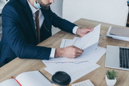 cropped view of businessman looking at papers with graphs and charts while sitting at workplace