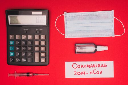 Top view of medical mask, calculator near syringe and card with coronavirus 2019-nCov lettering on red backgroundの写真素材
