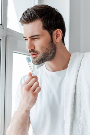 Bearded man holding towel and toothbrush while looking at window at homeの写真素材