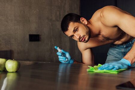 selective focus of muscular man in rubber gloves holding rag and bottle with antiseptic near apples on tableの写真素材