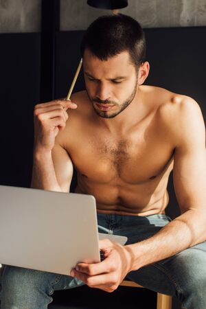 handsome and shirtless freelancer holding pencil and looking at laptopの写真素材